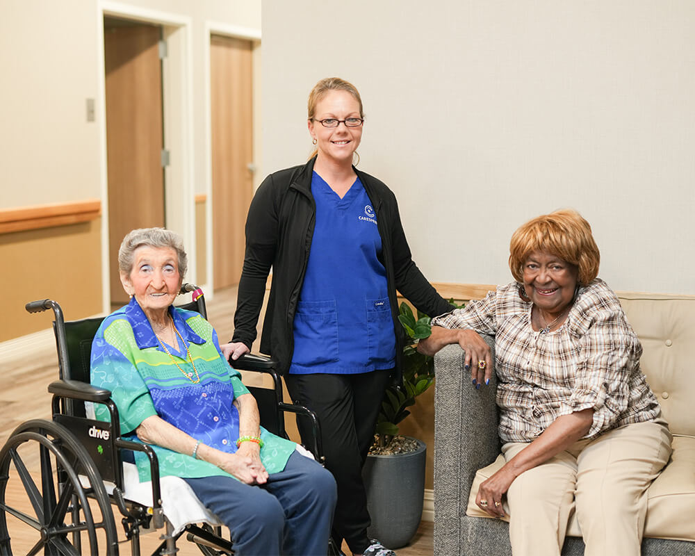Caregiver with two seniors, one seated in a wheelchair and one on a couch, smiling indoors.