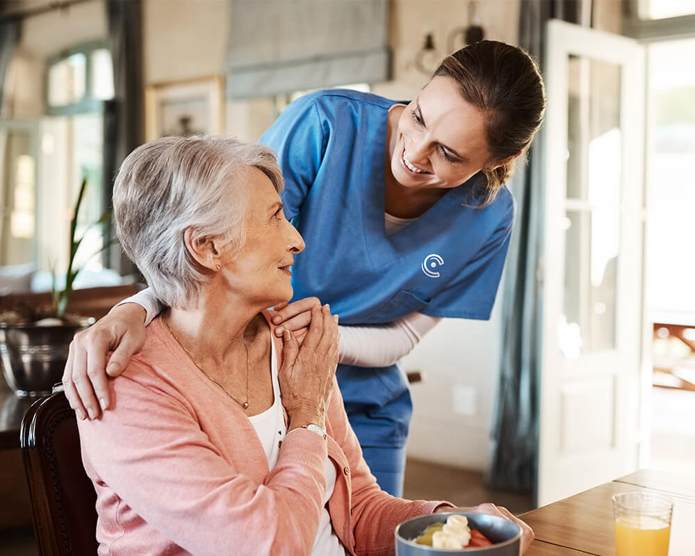 Caregiver smiling at senior woman holding a bowl of fruit in a cozy living space.
