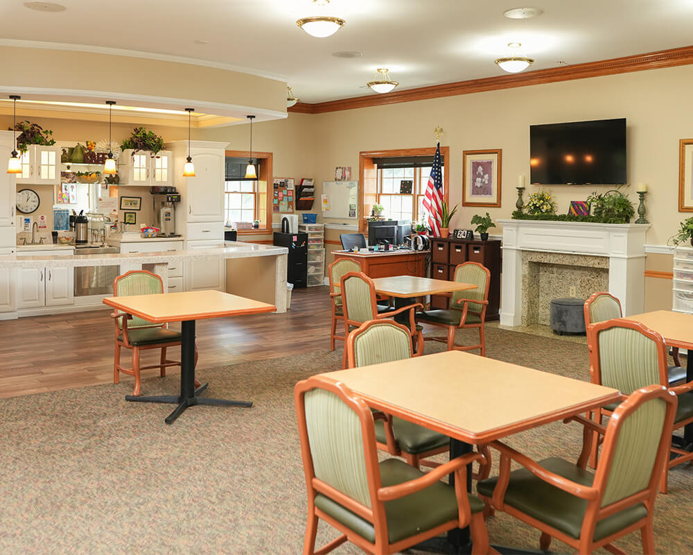 Warm community dining area with tables, chairs, and a kitchen in a senior living facility.