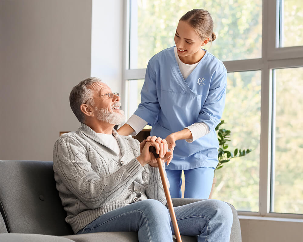 Caregiver assists smiling senior man with a cane by a large window.
