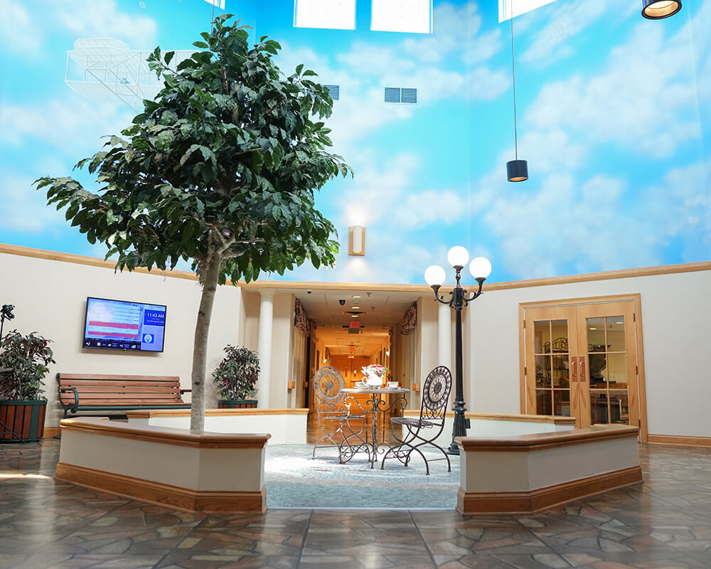 Indoor courtyard with seating, potted trees, and sky-themed ceiling in a residential facility.