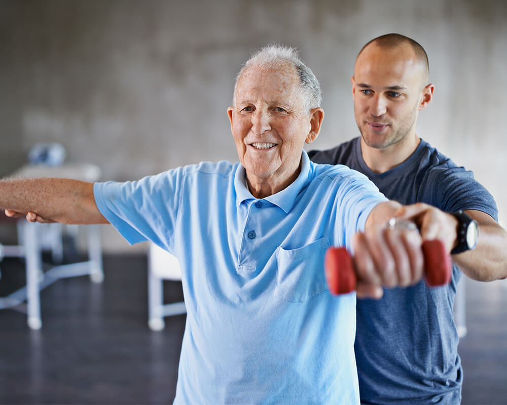 Elderly man exercises with assistance from a caregiver in a bright room.