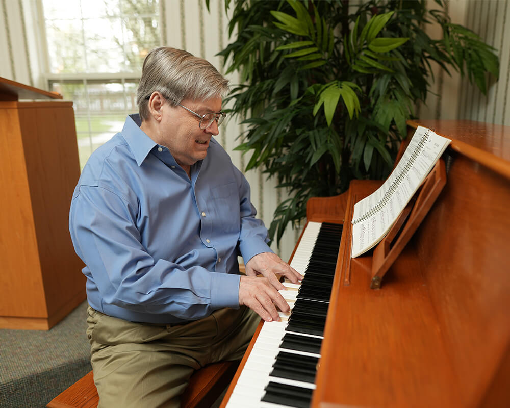 Older man playing piano in a living room with a large plant nearby.