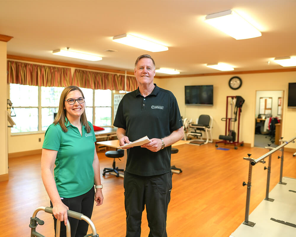Two staff members in a well-lit exercise room with therapy equipment.
