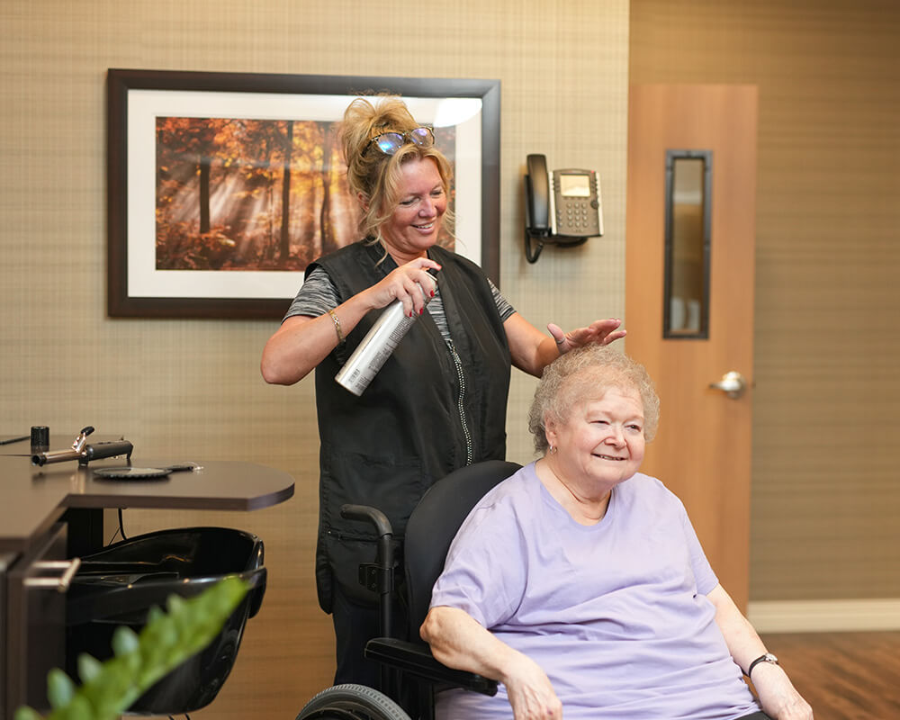 Woman in salon applying hairspray to seated elderly woman in purple shirt.