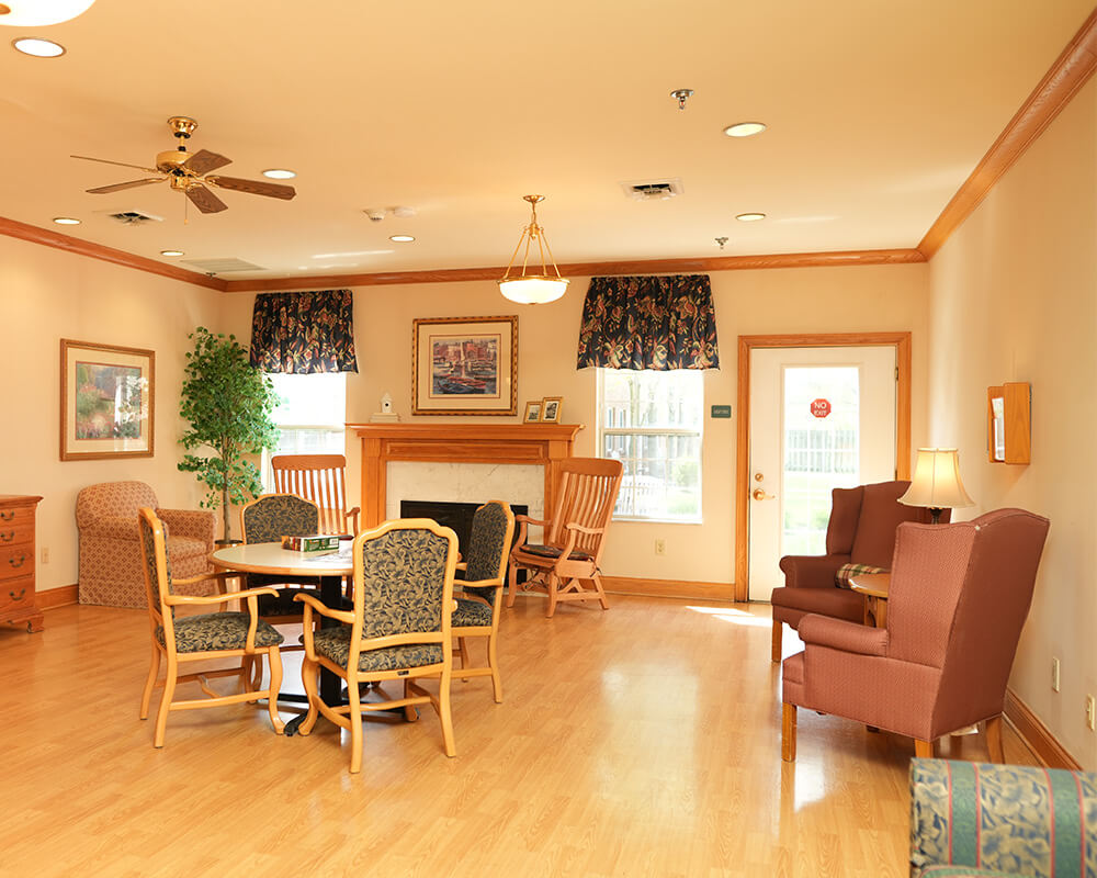 Cozy living area with chairs, table, lamp, and fireplace in a warm, sunlit room.