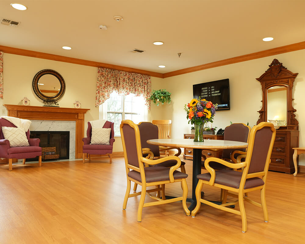 Cozy community space with round table, chairs, and fireplace in a senior living facility.