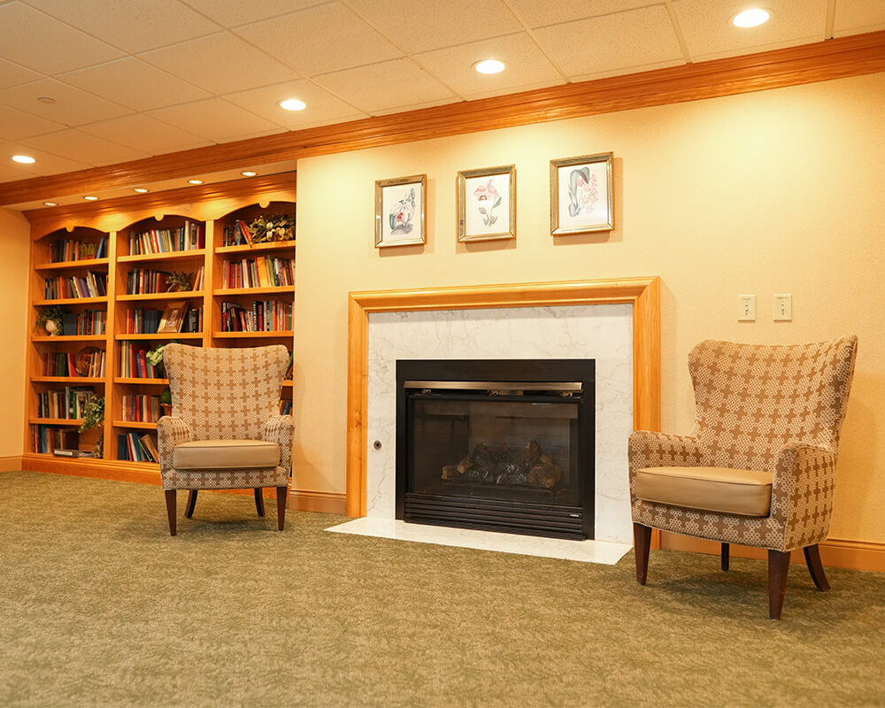 Cozy room with fireplace, two armchairs, bookshelves, and art in a community setting.