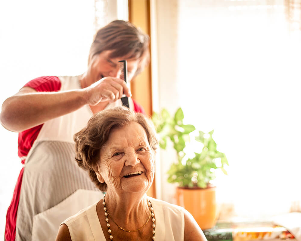 Elderly woman smiling while receiving hair care at a senior living community.