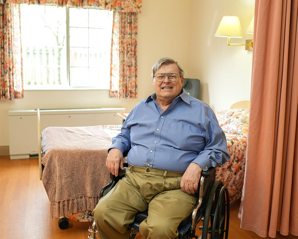 Man in wheelchair smiling in a bright, cozy living unit with floral decor.