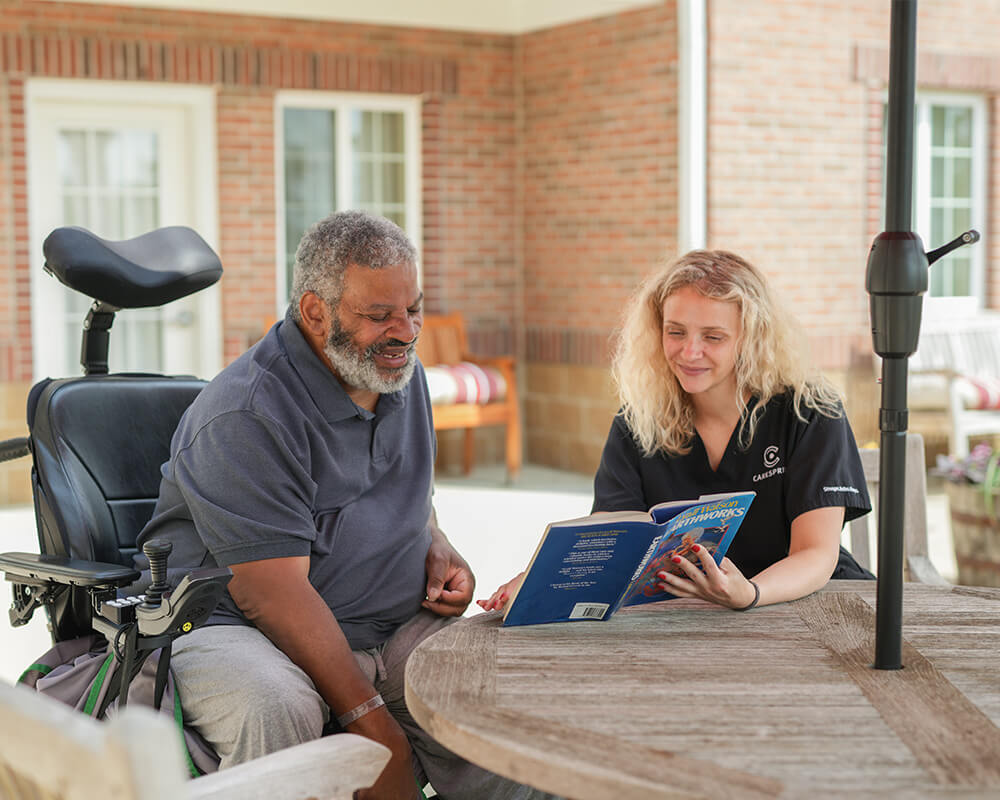 Elderly man in wheelchair enjoys book with caregiver at senior living community patio.