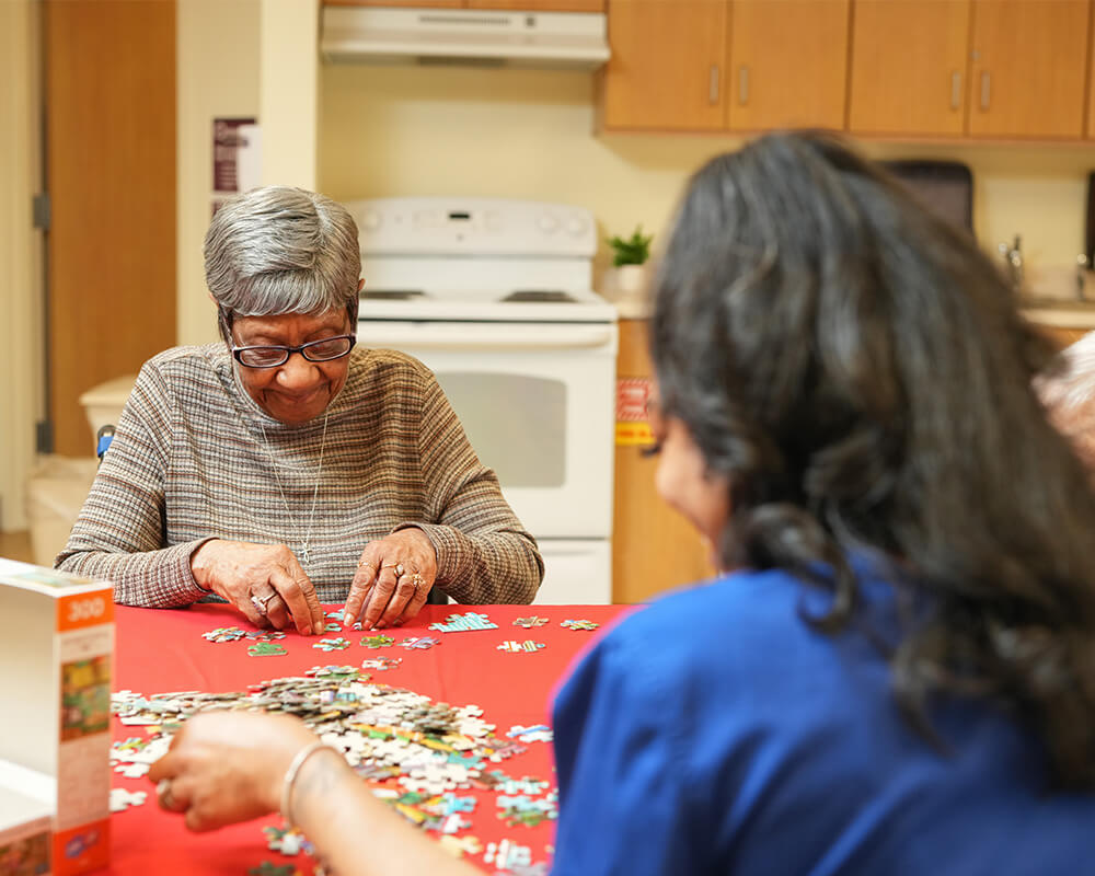 Two seniors assemble a jigsaw puzzle on a red table in a communal kitchen area.