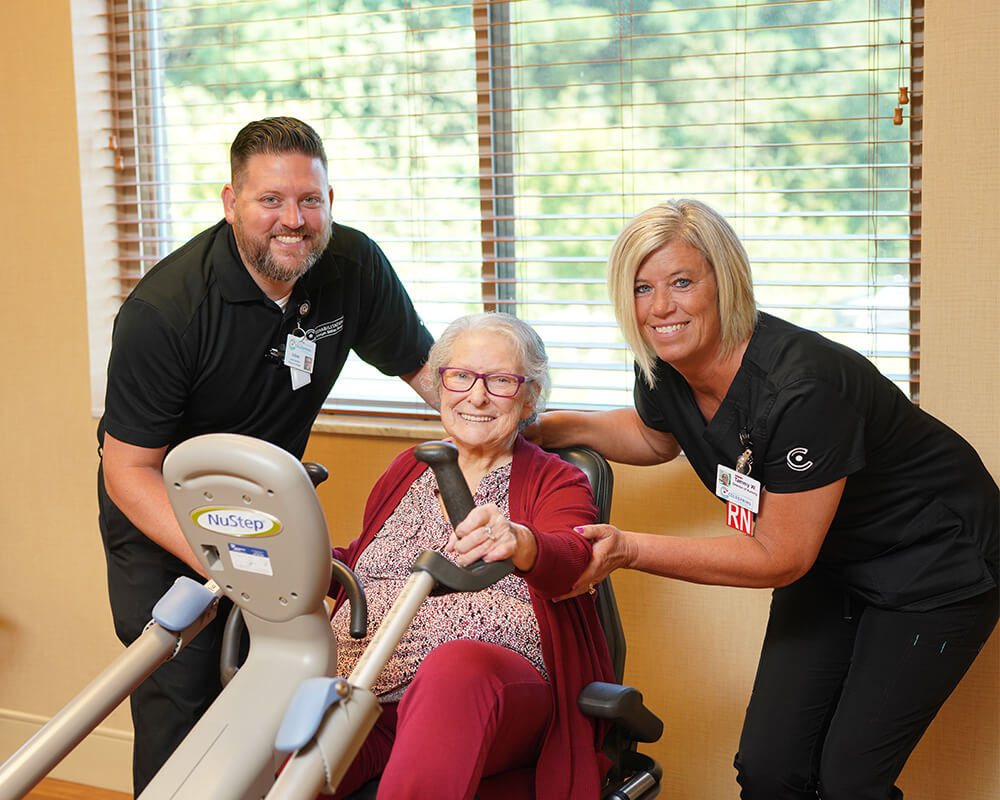 Senior woman using exercise equipment with two supportive staff members beside her.