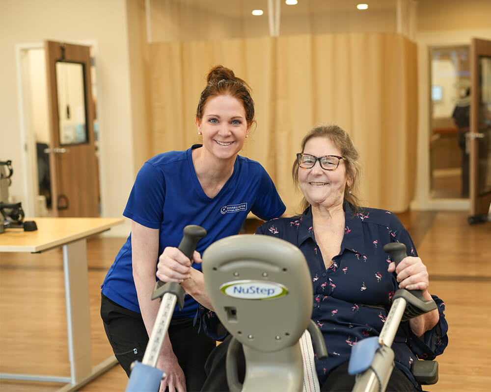 Smiling caregiver assists senior woman using exercise equipment indoors.