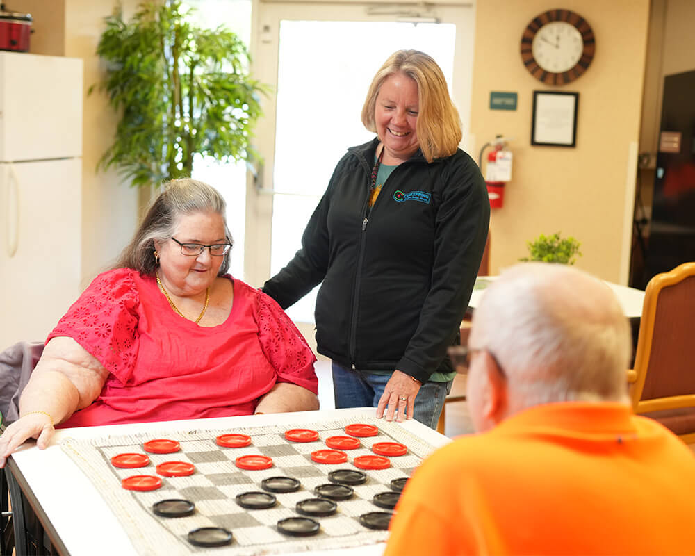 Residents playing checkers with a staff member in a senior living community common area.