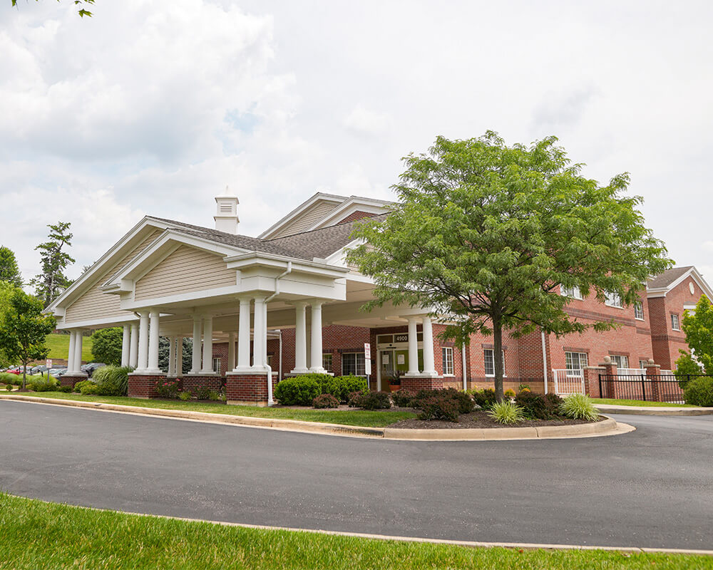 Brick and white building in a green landscape with a tree and driveway.