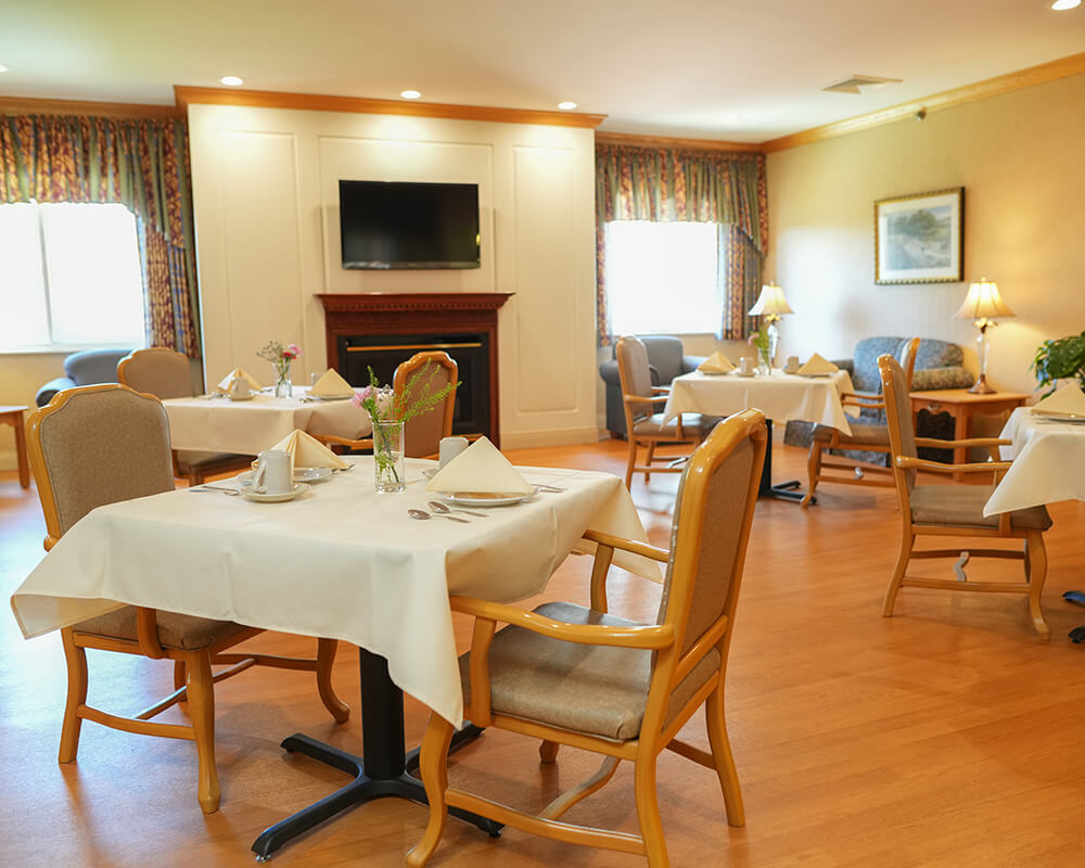 Cozy dining room in a senior living community with tables, chairs, and a TV on the wall.