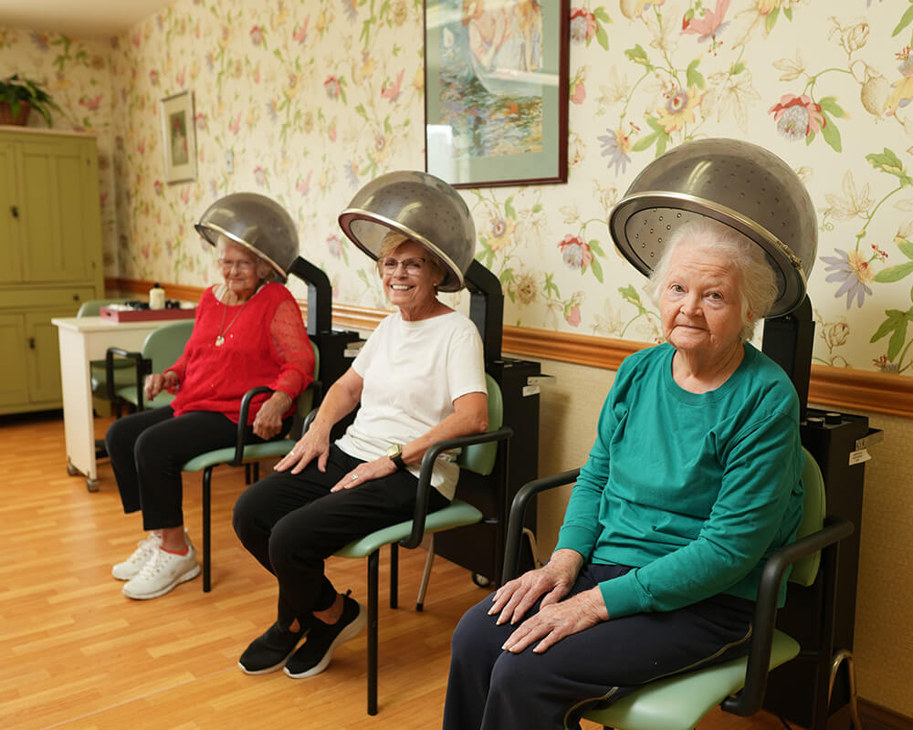 Three senior women enjoy a salon day under hair dryers in a cozy living community.