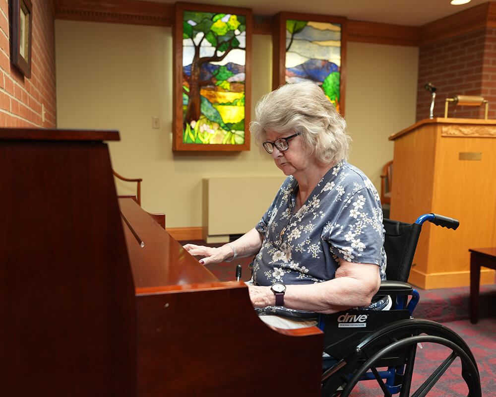 Senior woman in wheelchair playing a piano in a community room with stained glass art.