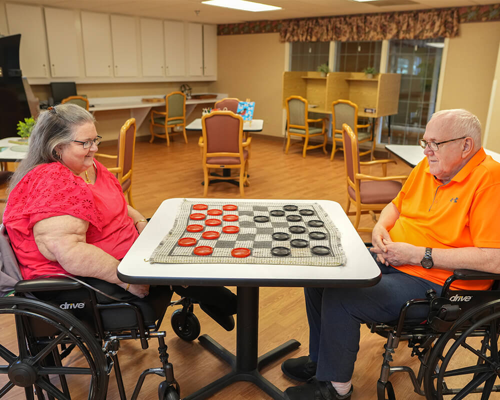 Two seniors in wheelchairs playing checkers in a communal room.