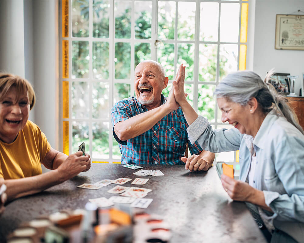 Group of seniors joyfully playing cards at a table in a sunlit room.