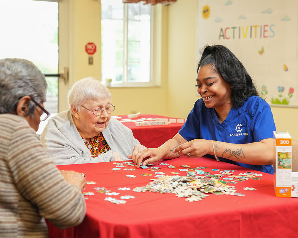 Caregiver assists seniors in assembling a puzzle at a community activity table.