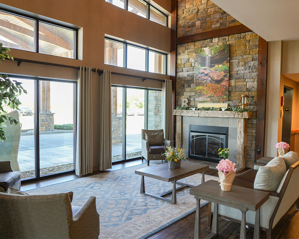 Spacious lounge area in a senior living community with large windows and a stone fireplace.