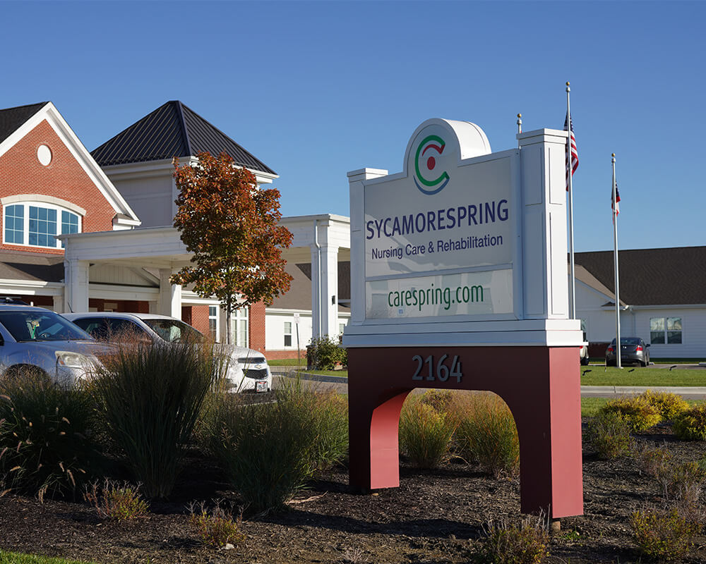 Sign for Sycamore Spring community with building and cars in the background.
