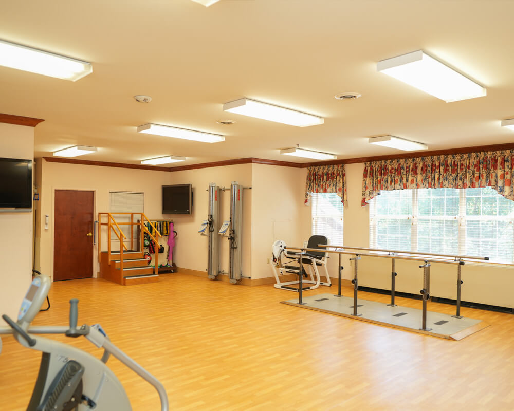 Well-lit exercise room with equipment and large windows in a living community.