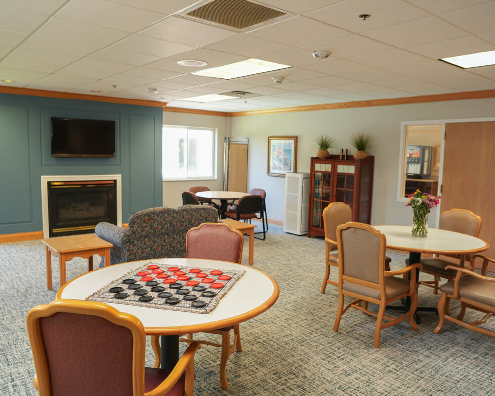 Cozy community room with tables, chairs, fireplace, and a checkers game setup.