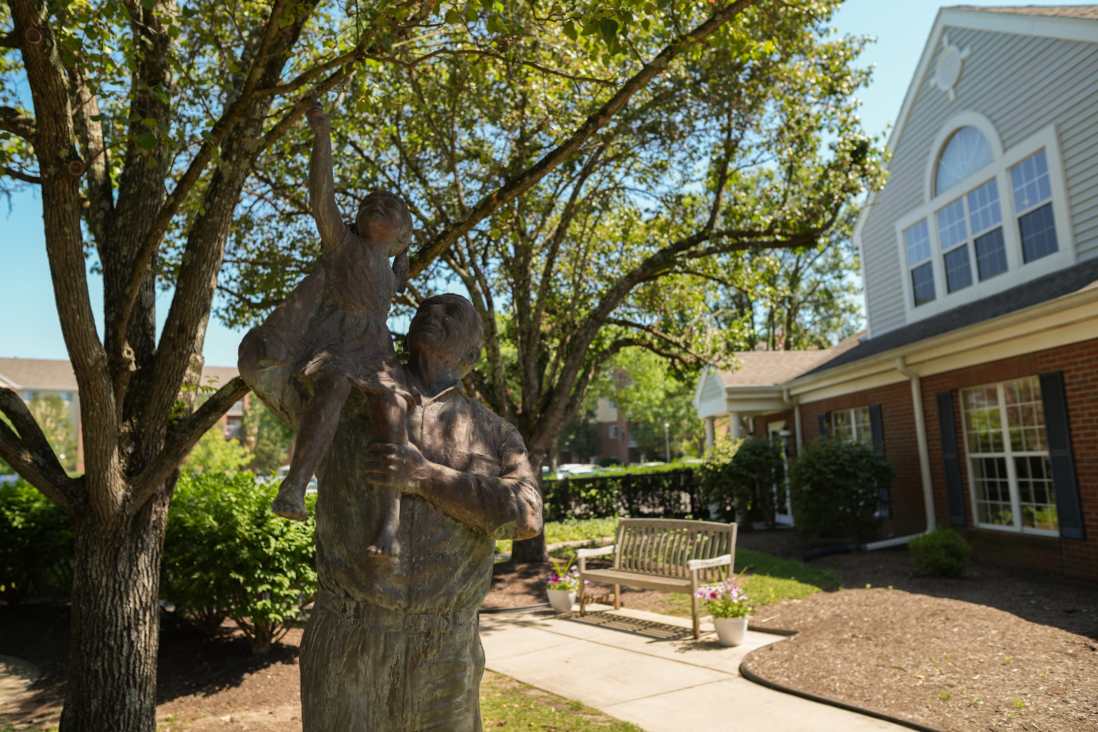 Bronze statue of man lifting child under trees near senior living units.