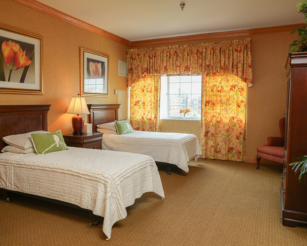 Cozy senior living unit with twin beds, floral curtains, and warm lighting.