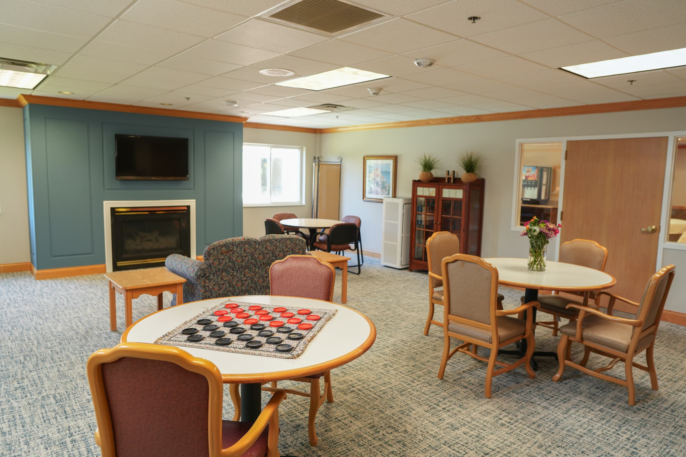 Communal lounge with tables, chairs, TV, fireplace, and checkers in a senior living building.