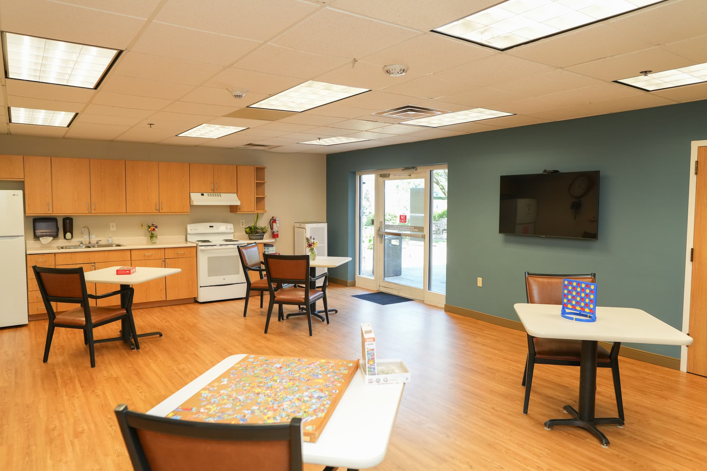 Community room with kitchen, tables, and games in a senior living building.