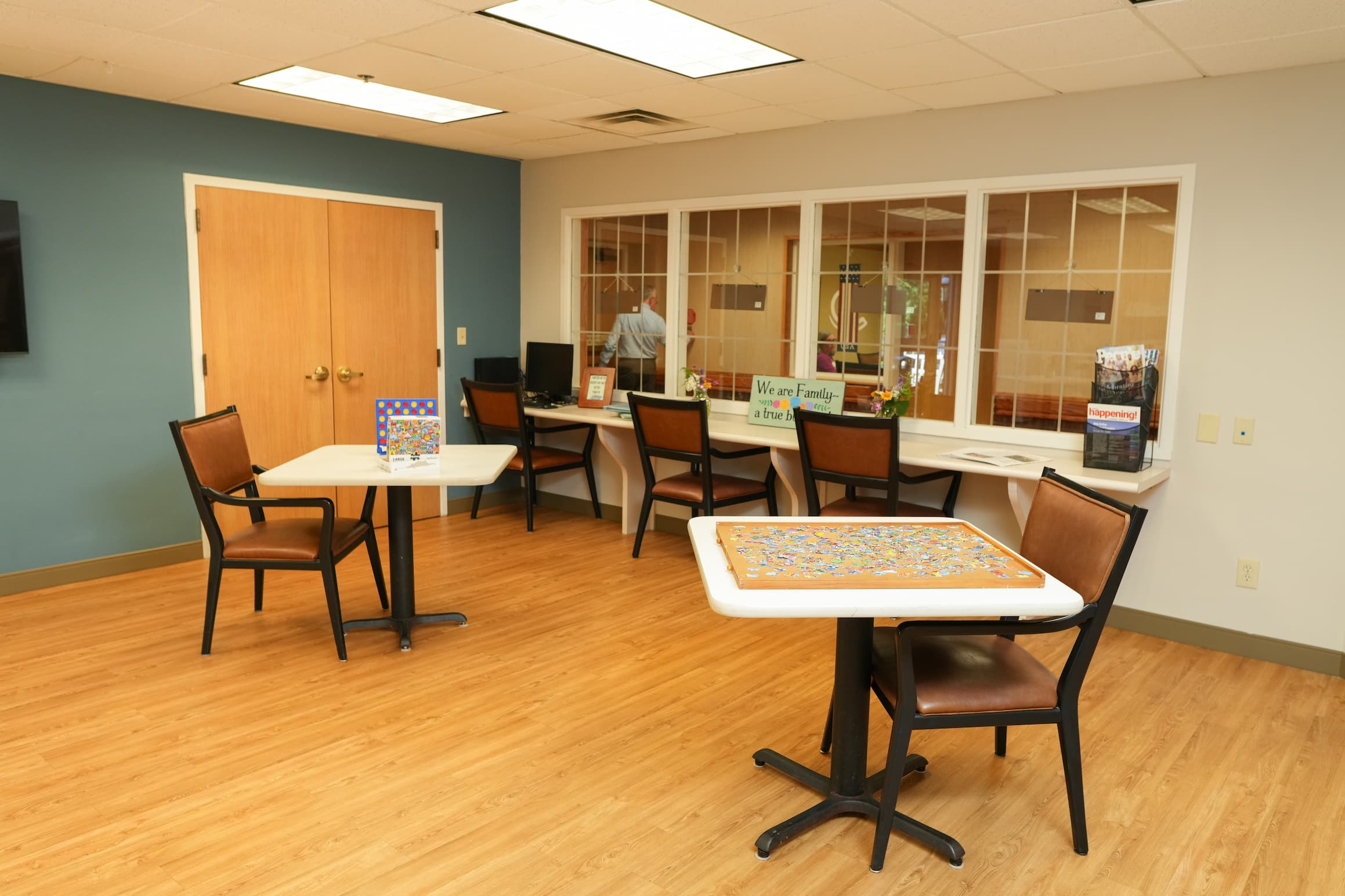 Community room with tables, chairs, and a puzzle on a table in a senior living building.