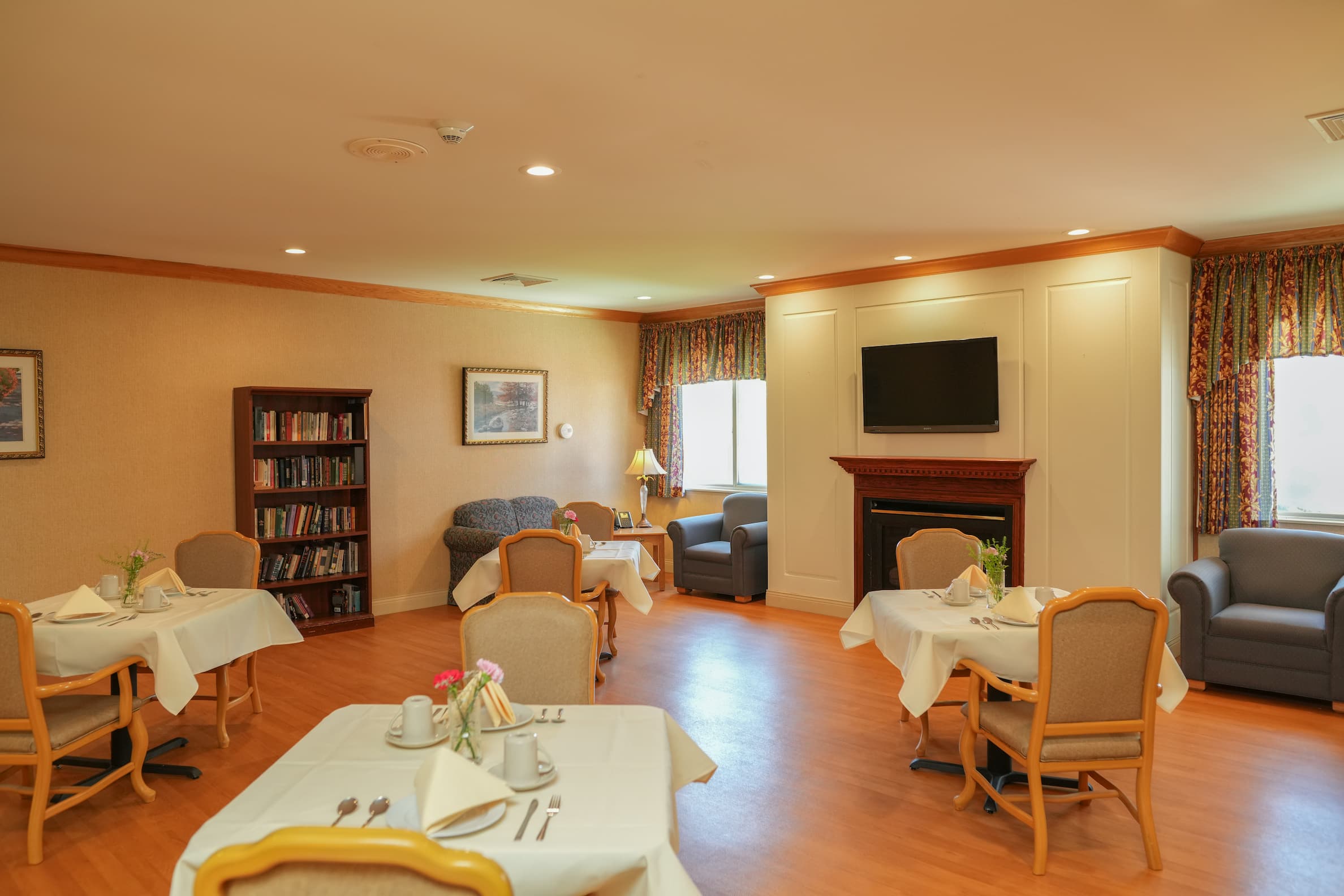 Cozy community room with tables, chairs, and a fireplace in a senior living facility.