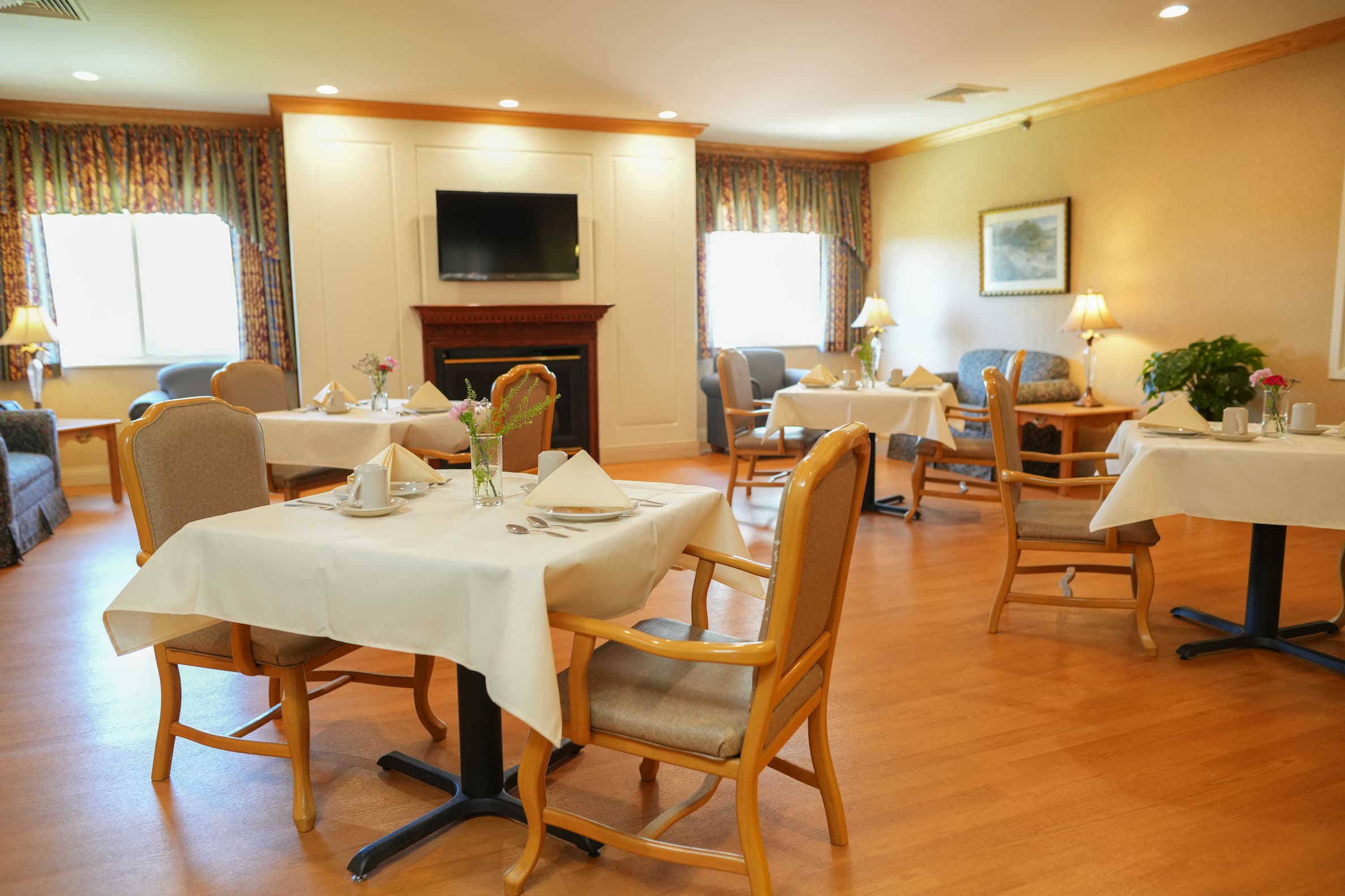 Dining area in community with tables set for four, chairs, a fireplace, and a TV.
