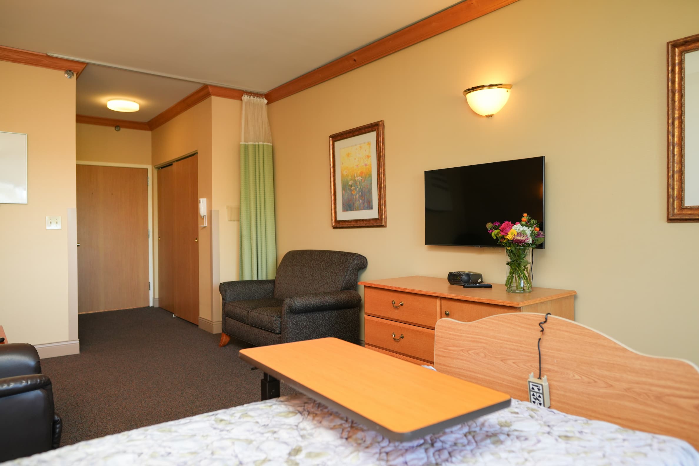 Cozy living space with a bed, armchair, TV, and flowers on a dresser.
