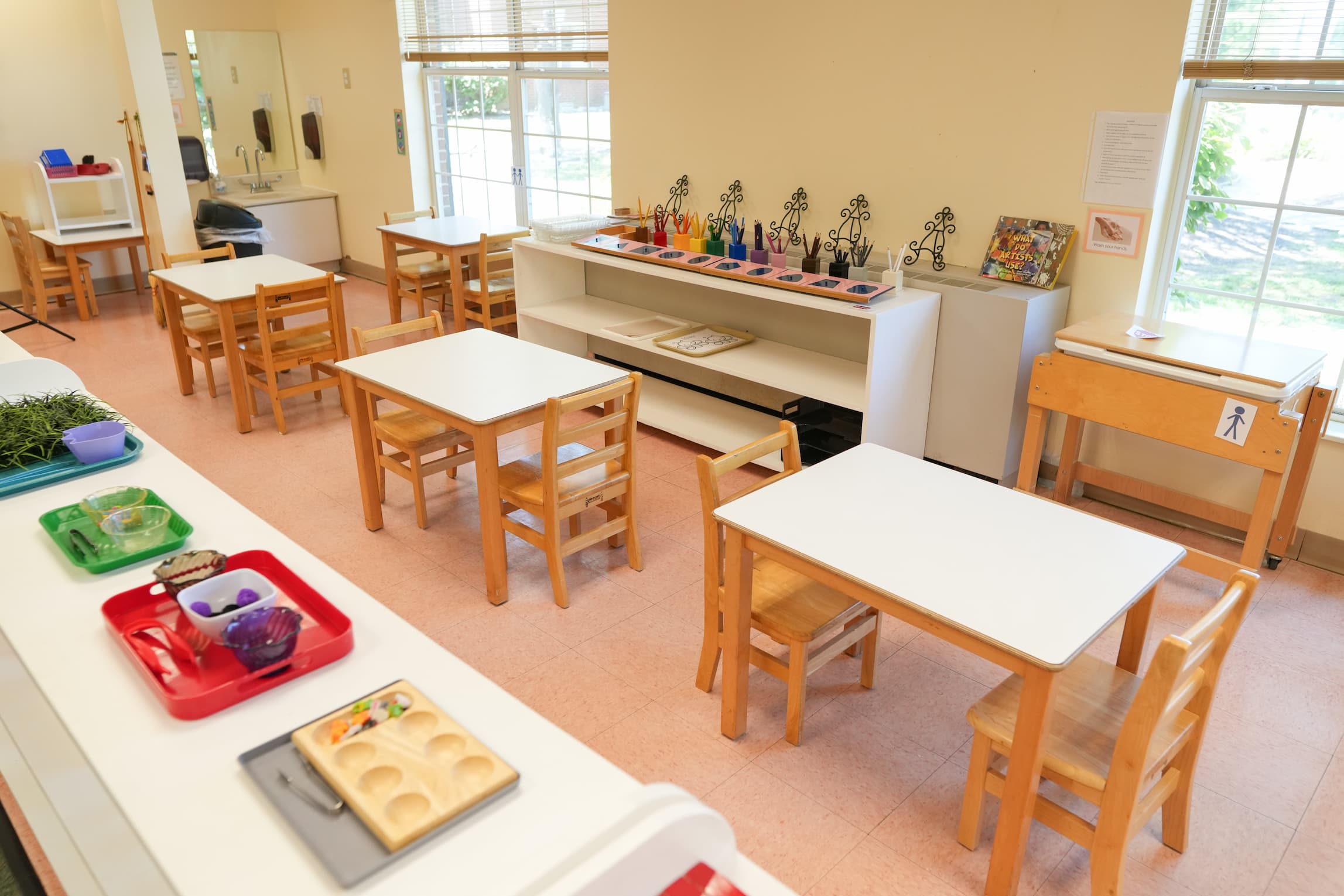 Bright classroom with small tables, chairs, craft supplies, and large windows.