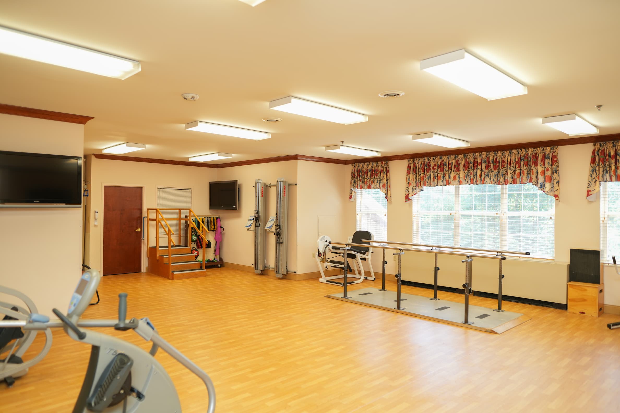 Exercise room with equipment and large windows in senior living community.