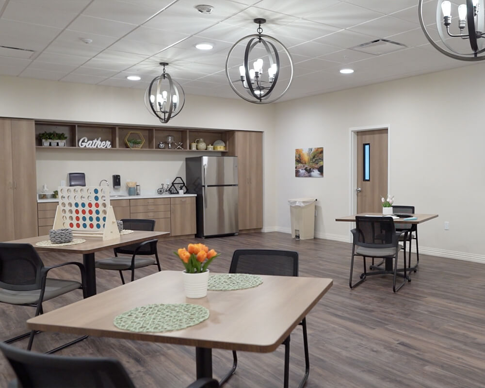 Cozy activity room with tables, chairs, chandelier lighting, and kitchen area.