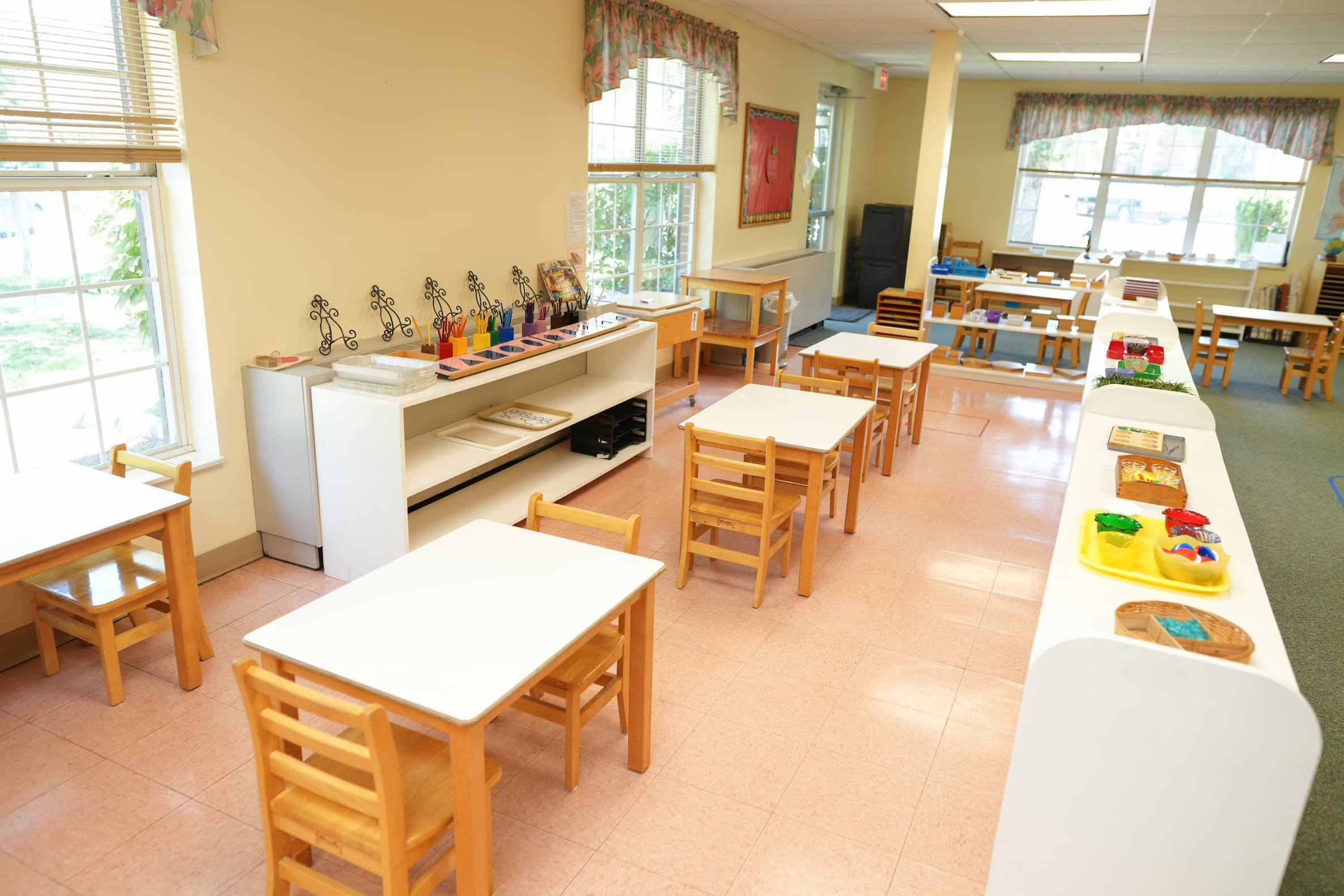 Bright classroom with small wooden tables and chairs, art supplies, and natural light.