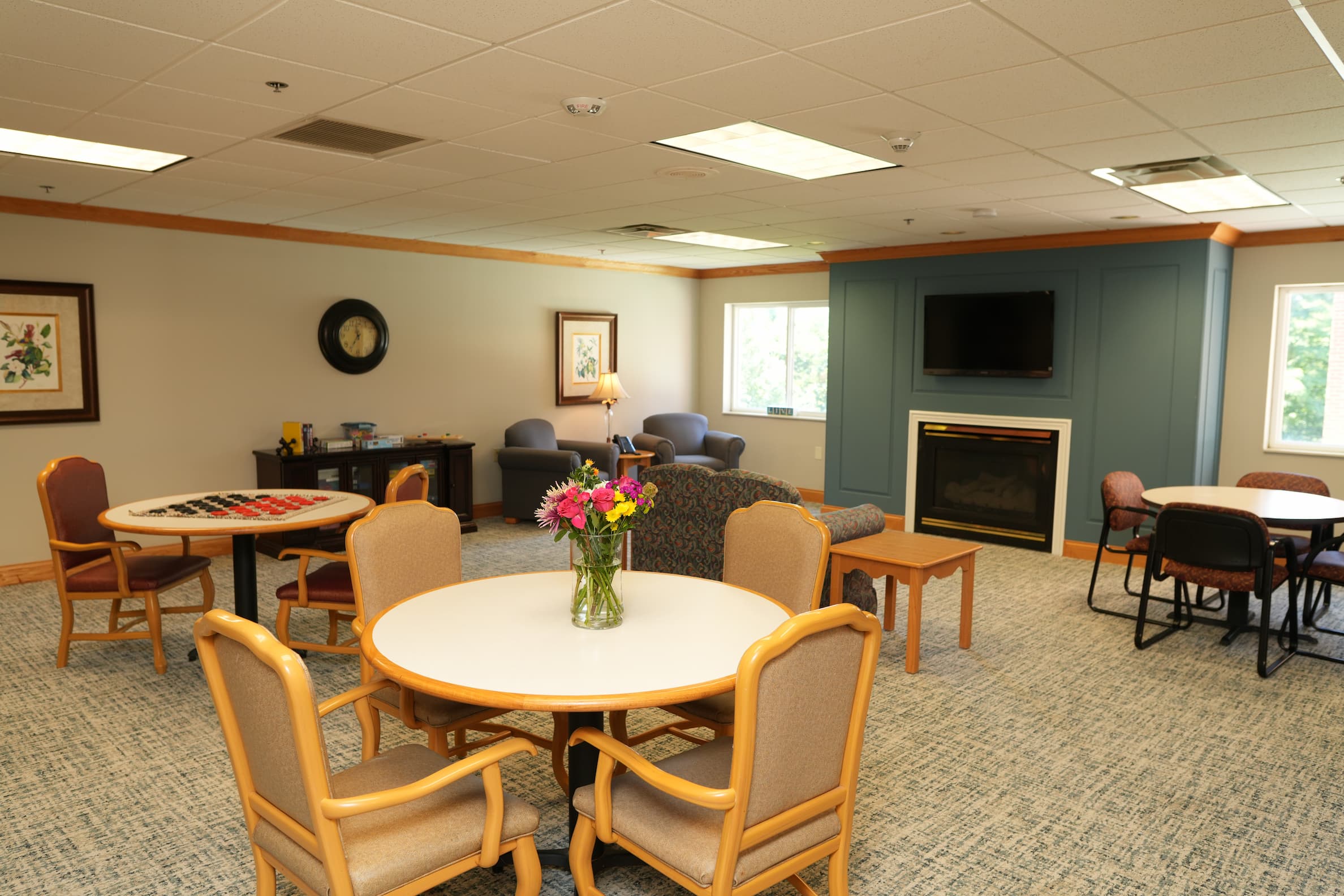 Common area with tables, chairs, a fireplace, and a vase of flowers in a senior living unit.