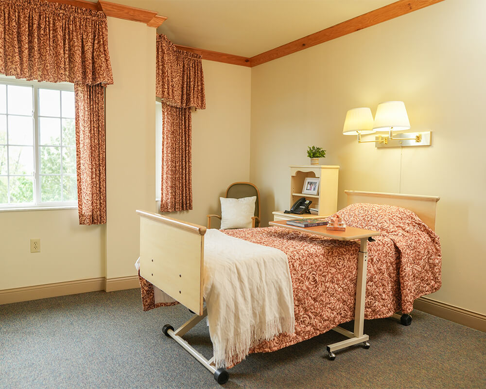Comfortable senior living unit with bed, window, and bedside table with books.