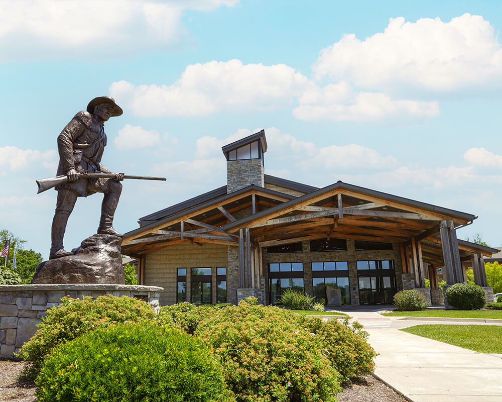 Statue of pioneer outside modern building with landscaped garden and blue sky.