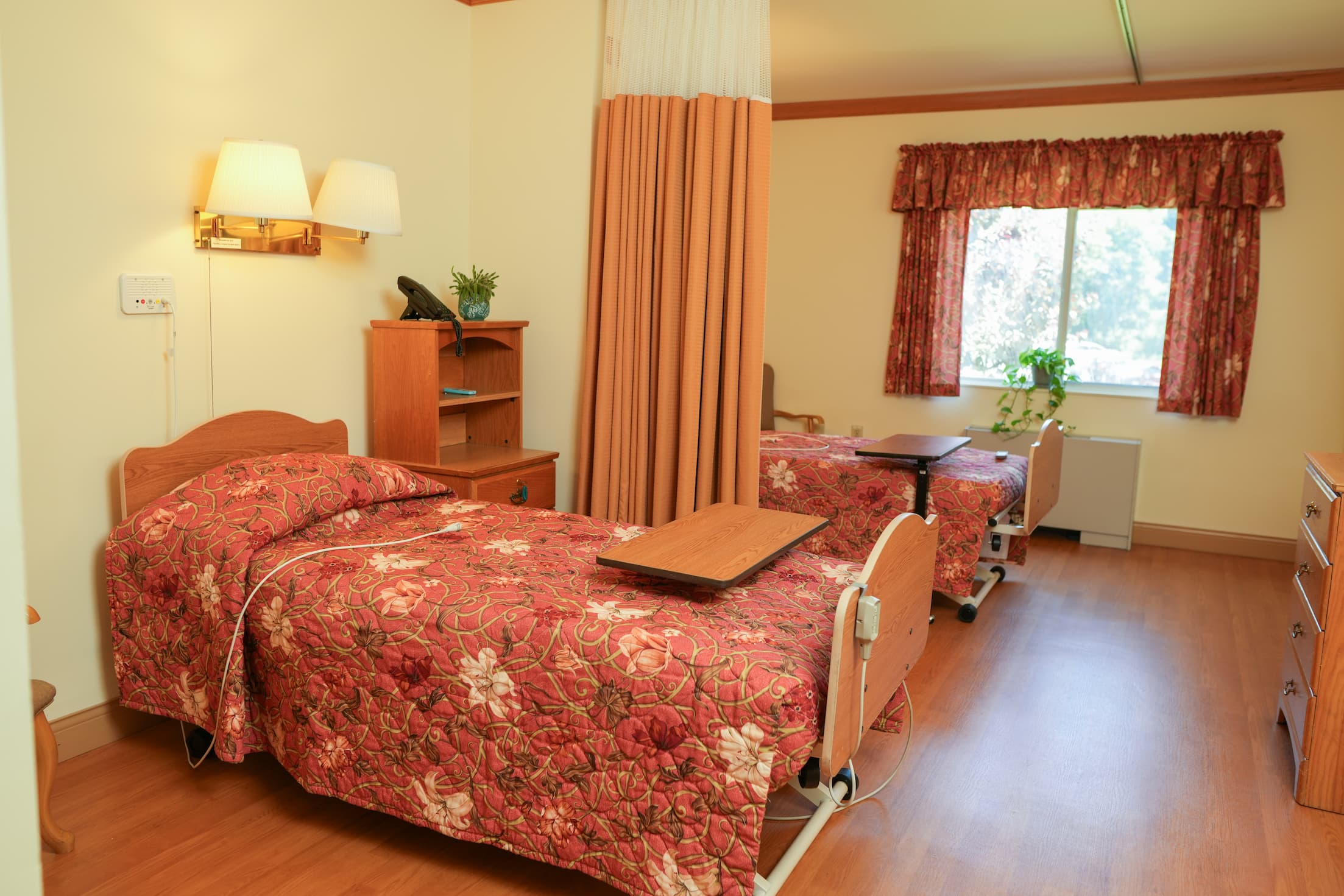 Senior living unit with two beds, floral bedding, wooden furniture, and a large window.